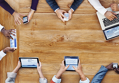 wooden table with business people hands holding various devices: cell phones, tablets, typing on laptop